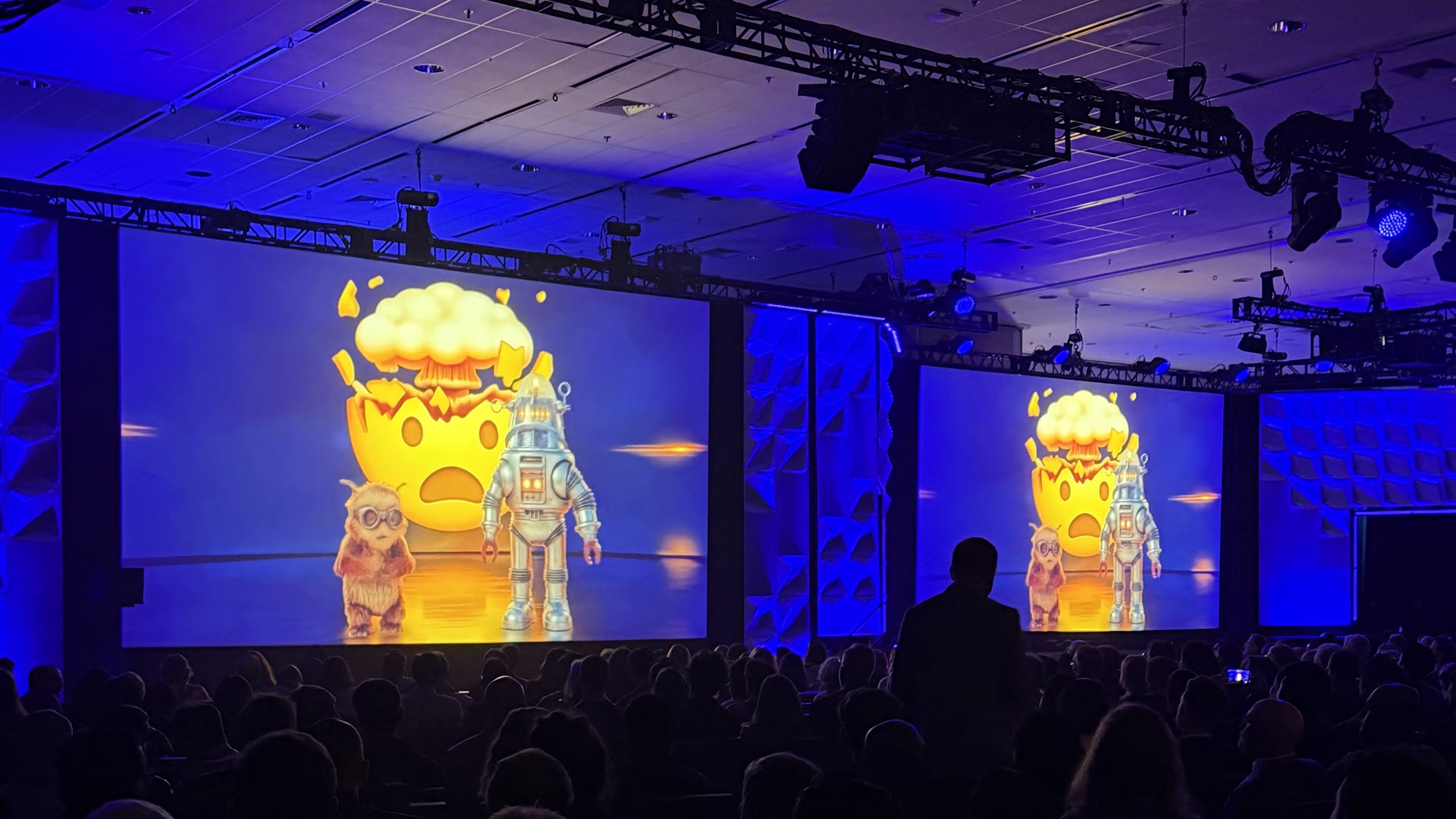 A packed conference keynote bathed in blue light, its giant screens showing an exploding-head emoji character flanked by a robot, towering over the audience