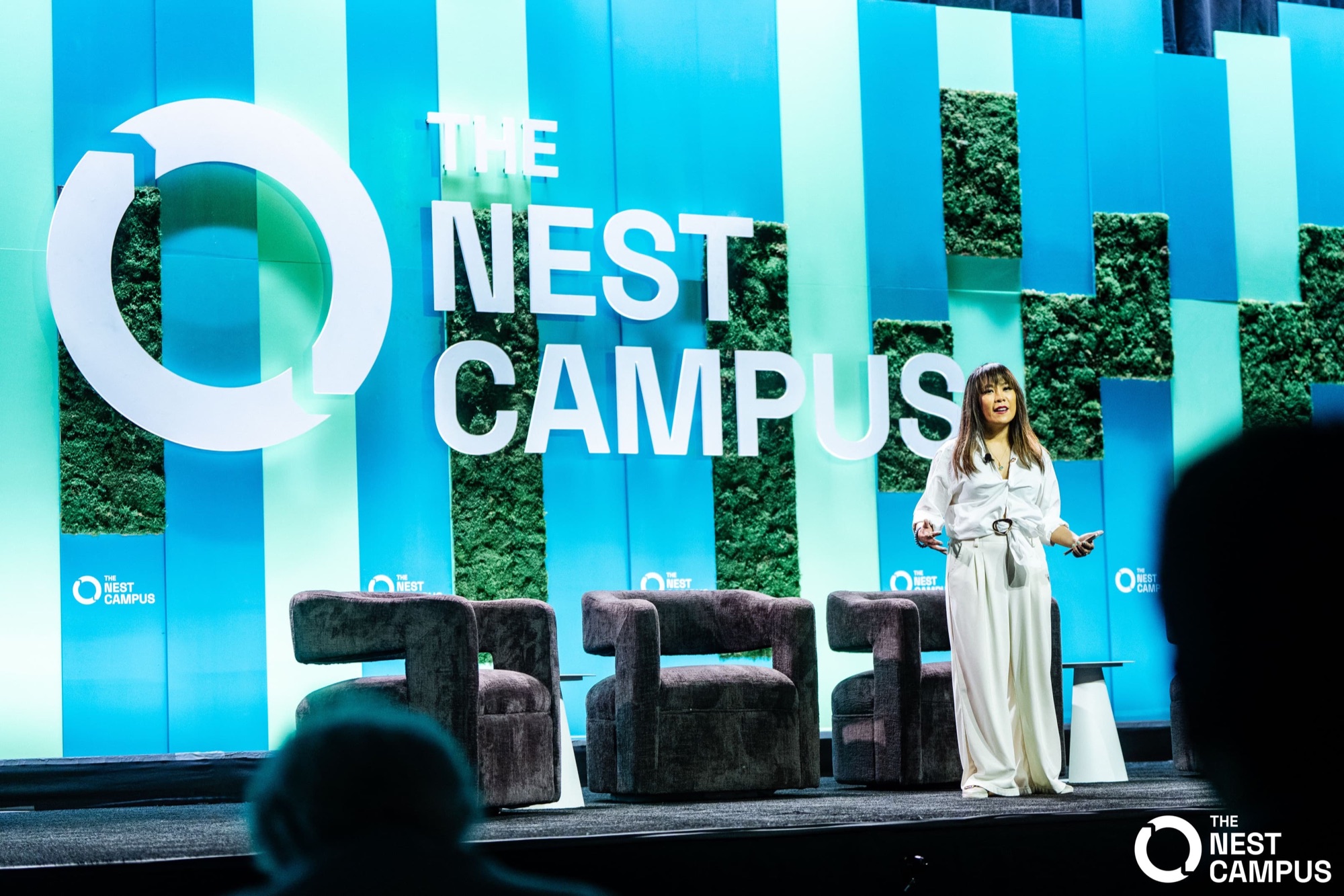 Kat Dunn speaking on the Main Stage at The Nest Campus, in front of the moss-and-teal Nest Campus backdrop
